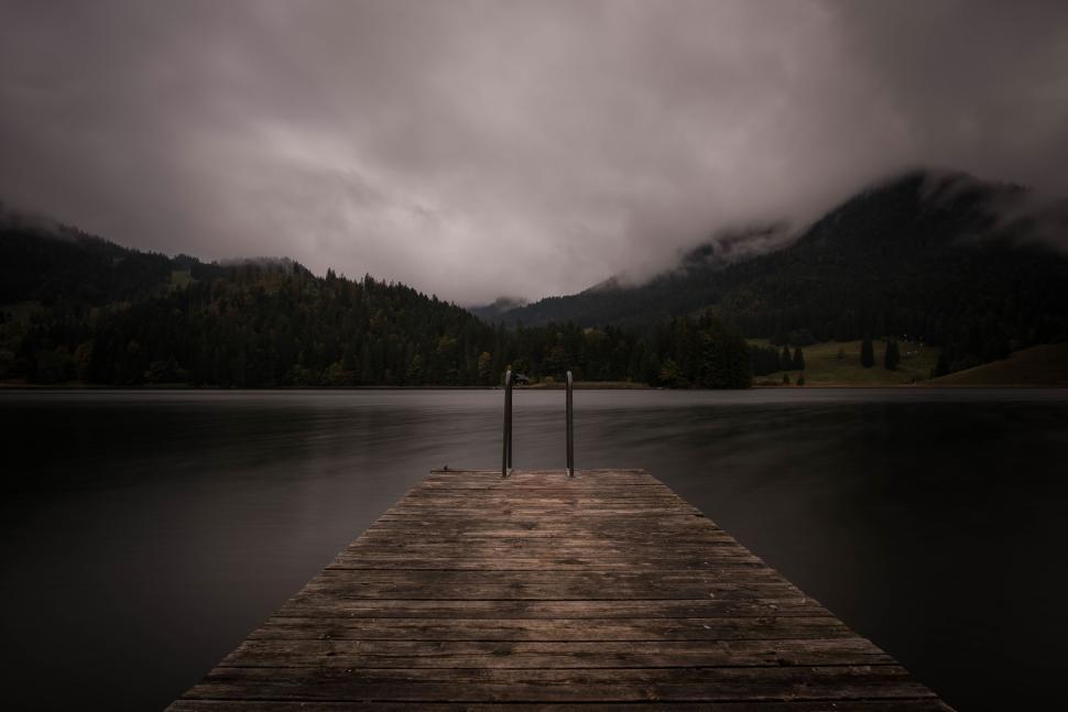 Free Image of Wooden dock leading into a still lake with cloudy sky 