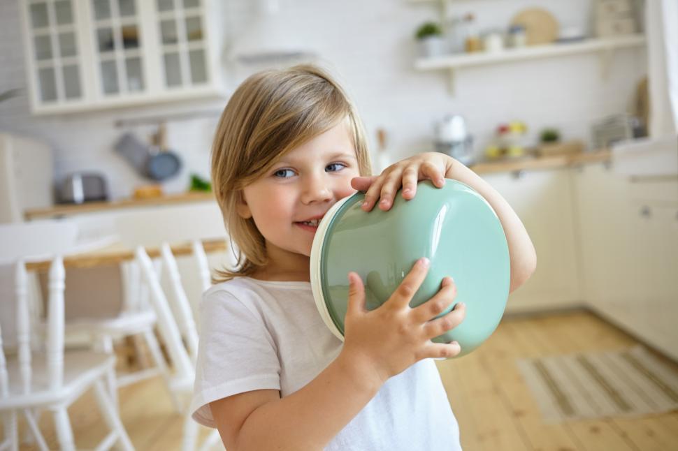 Free Image of Portrait of playful charming European female kid with fair hair holding saucepan, going to help mother to cook soup for dinner, standing against blurred kitchen interior background and smiling 