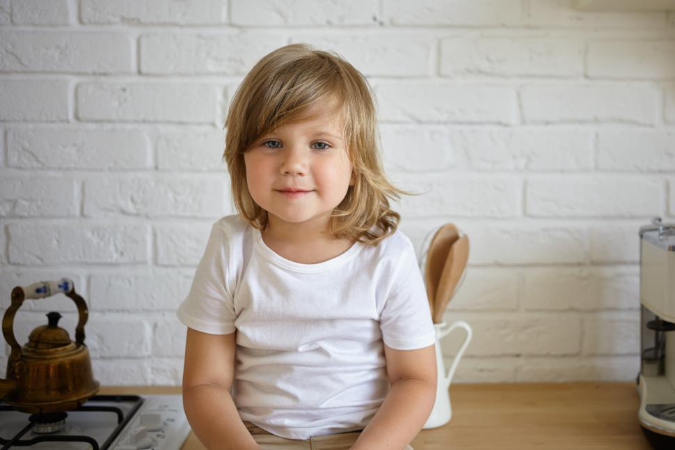Free Image of Indoor shot of little 5 year old baby girl dressed casually sitting on kitchen counter at white brick wall and looking at camera with charming smile. Happy carefree childhood and leisure concept 