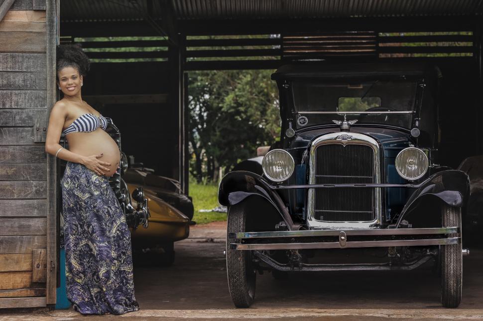 Free Image of Pregnant woman leaning on barn wall beside a vintage car. 