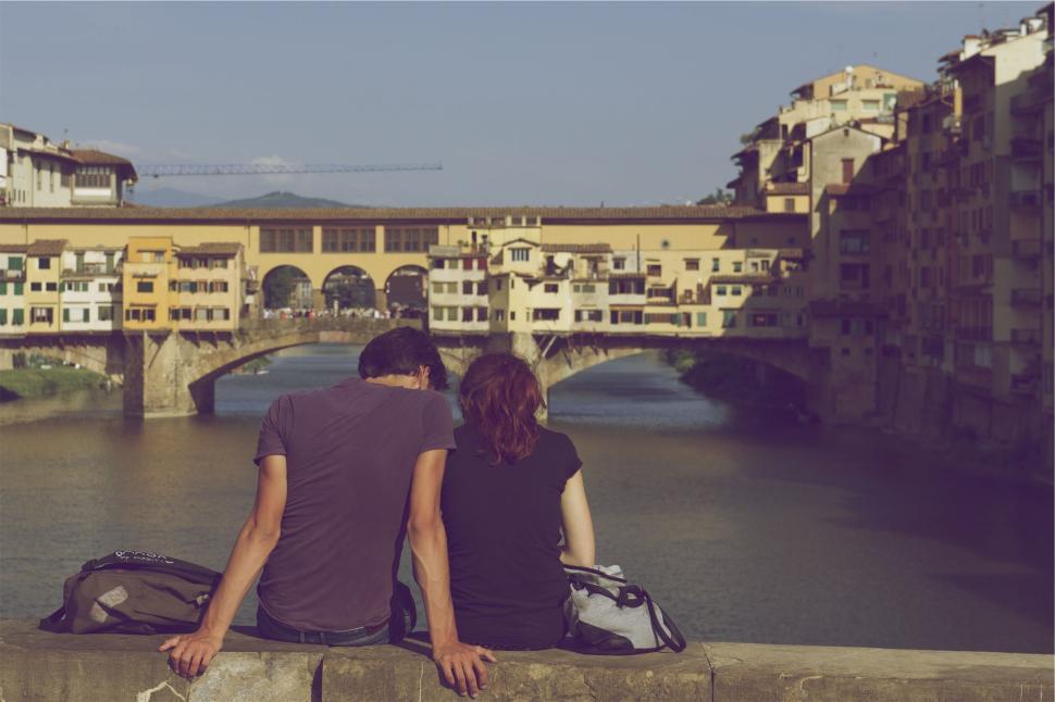 Free Image of Couple sitting on bridge railing overlooking river water 