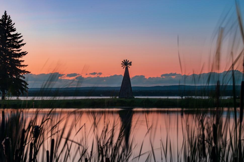 Free Image of Sunset by a windmill reflecting over calm lake waters 