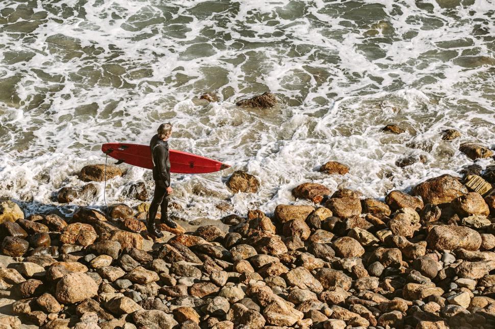 Free Image of Surfer in wetsuit standing on rocky beach with surfboard. 