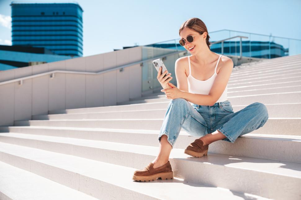 Free Image of Young woman enjoys a sunny day while texting on her phone in a modern urban setting 