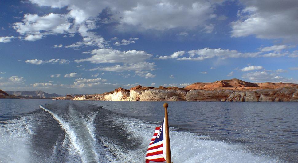Free Image of Vibrant lake landscape with boat trails and rocky cliffs under a blue sky 