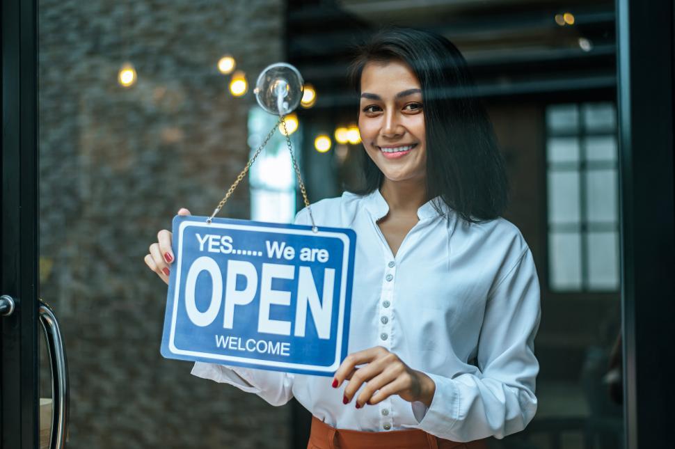 Free Image of woman stands and opens a wide sign through the shop window and smile. 