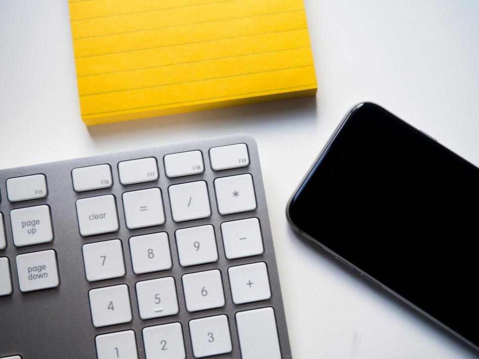 Free Image of Workspace setup with keyboard, smartphone, and yellow notepad on white desk 