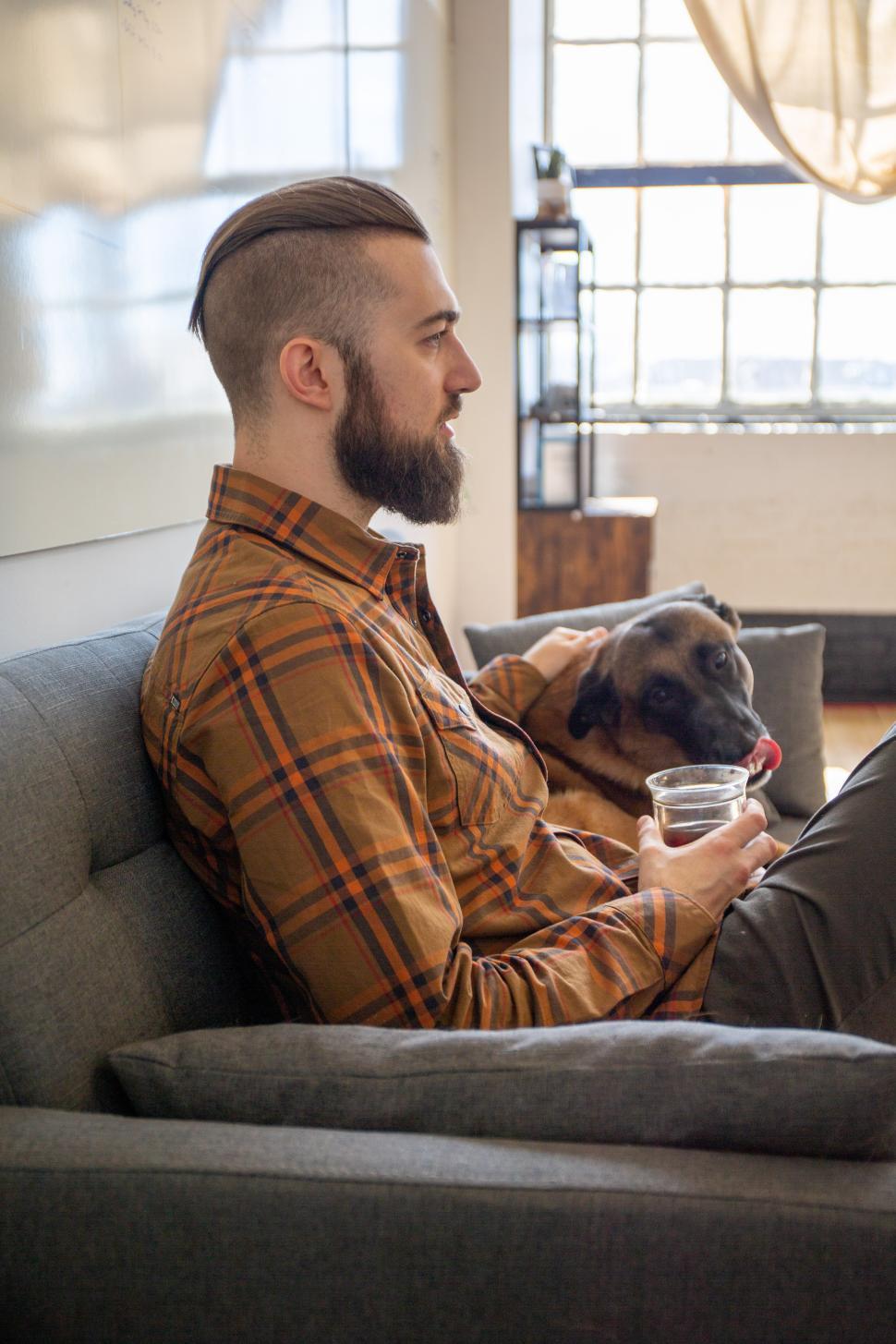 Free Image of Man relaxes on couch with dog and drink in cozy setting 