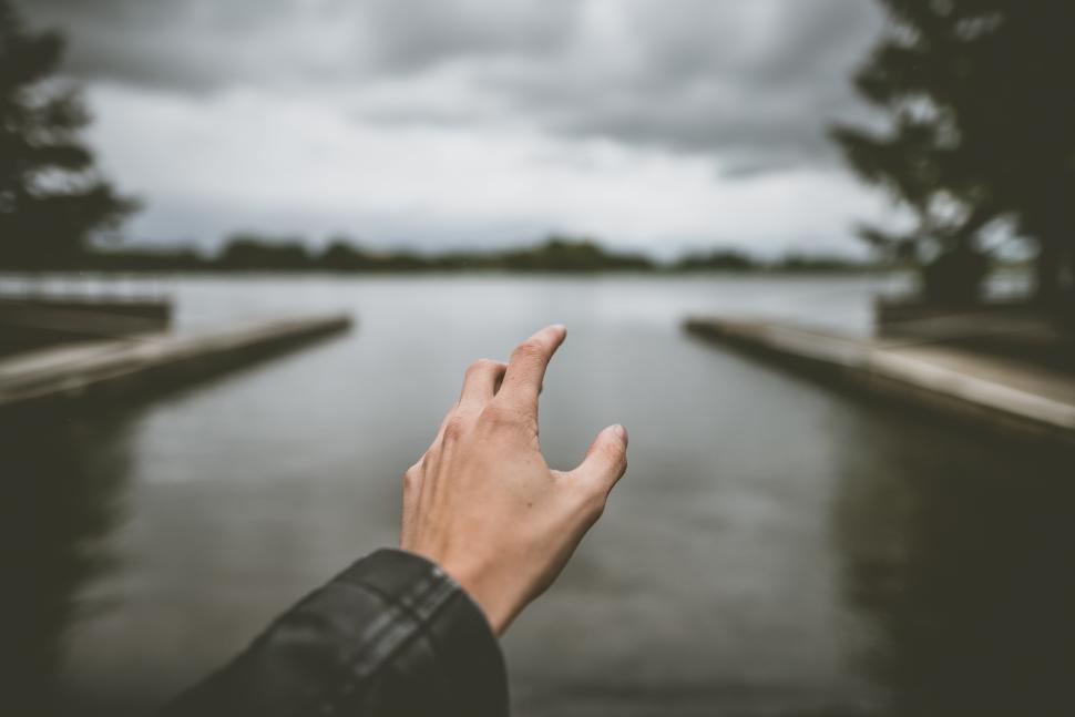 Free Image of Hand reaching out towards calm water on an overcast day by a lakeshore 