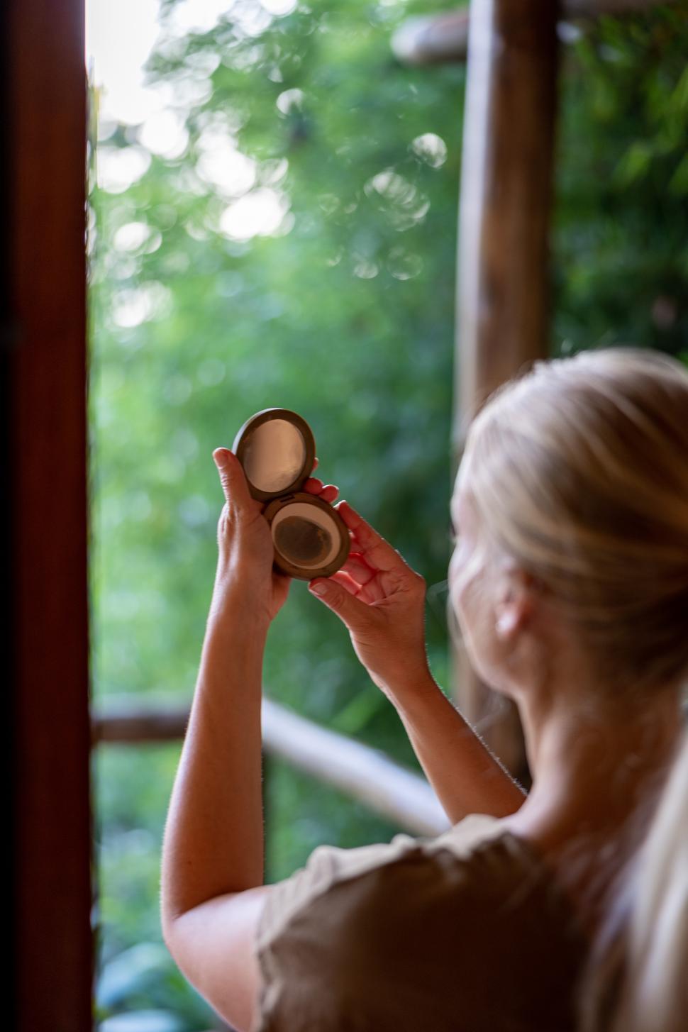 Free Image of Young woman examines compact mirror outdoors during sunny day 