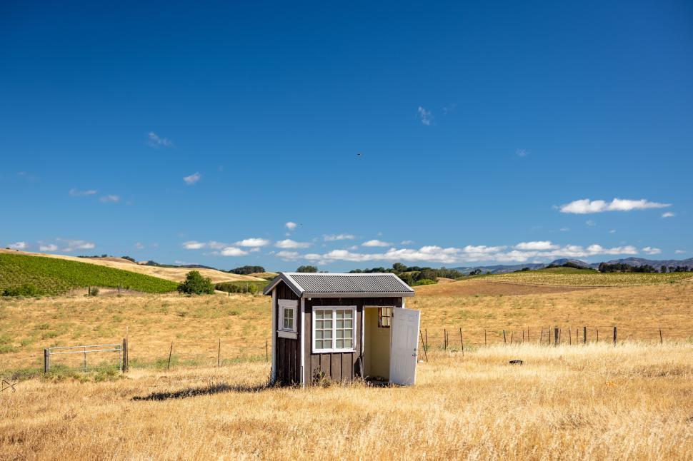 Free Image of Small wooden shack stands alone in a vast open field under blue sky 