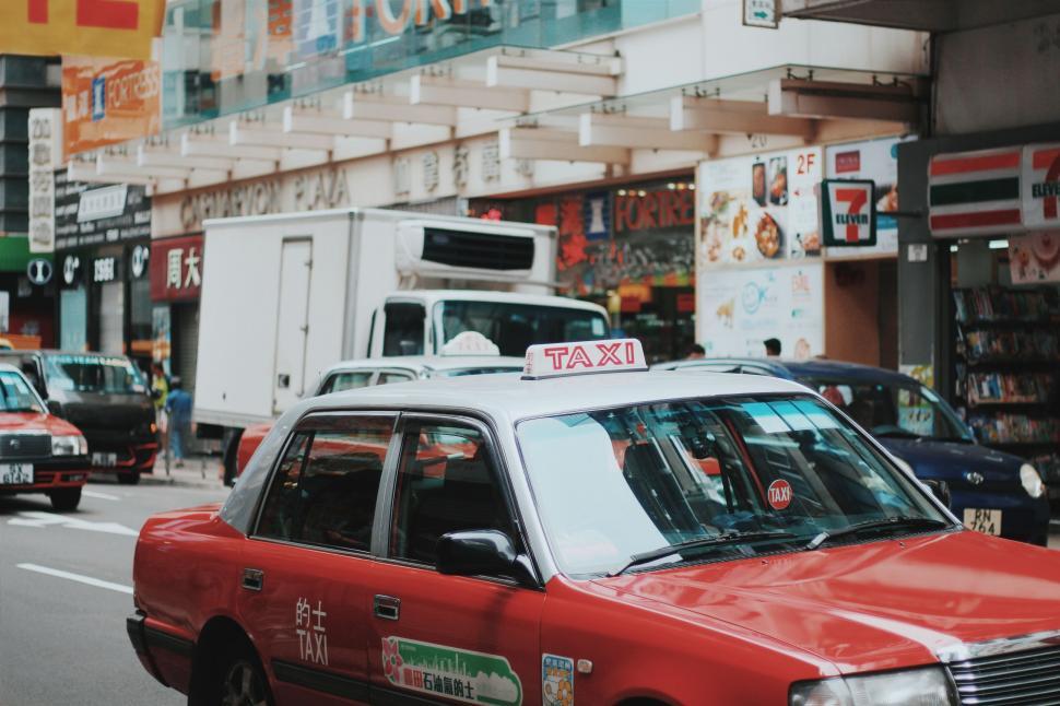 Free Image of Red Taxi Cab Driving Down Street Next to Tall Buildings 
