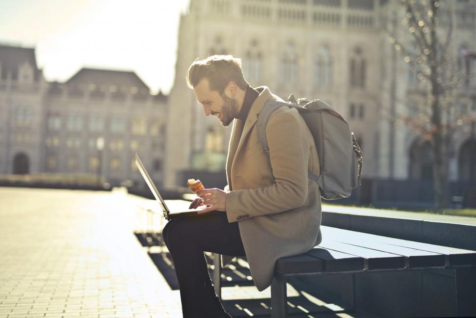 Free Image of Young Adult Man using a laptop while sitting on a bench during a 