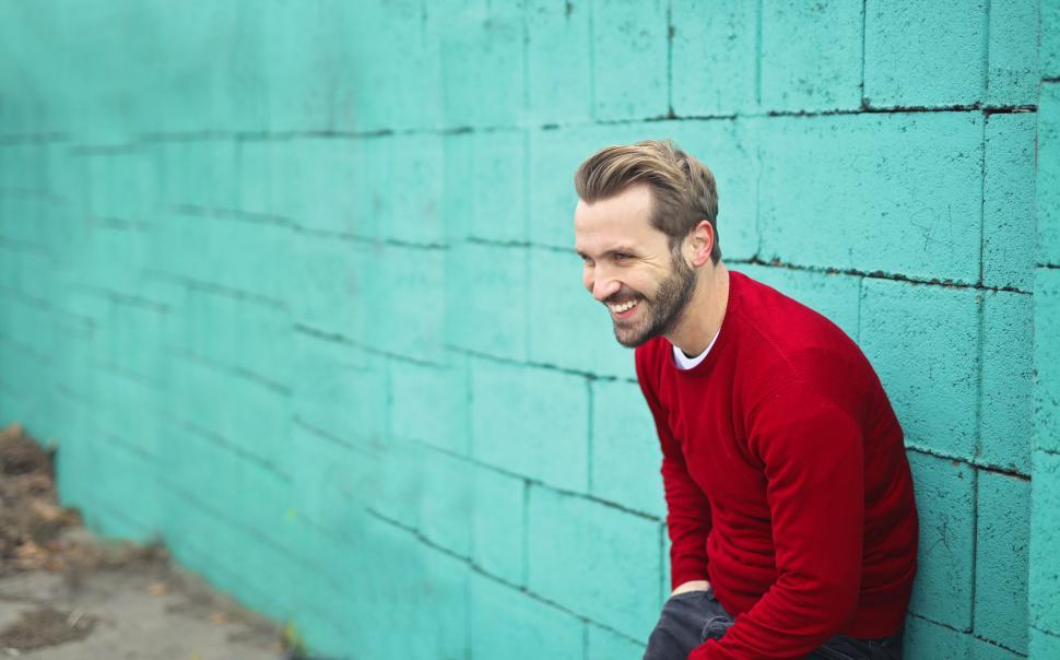 Free Image of Young Man Wearing a Red Sweater, Smiling and Leaning on a Blue W 