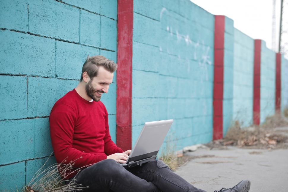 Free Image of A young bearded man sitting against a wall working 
