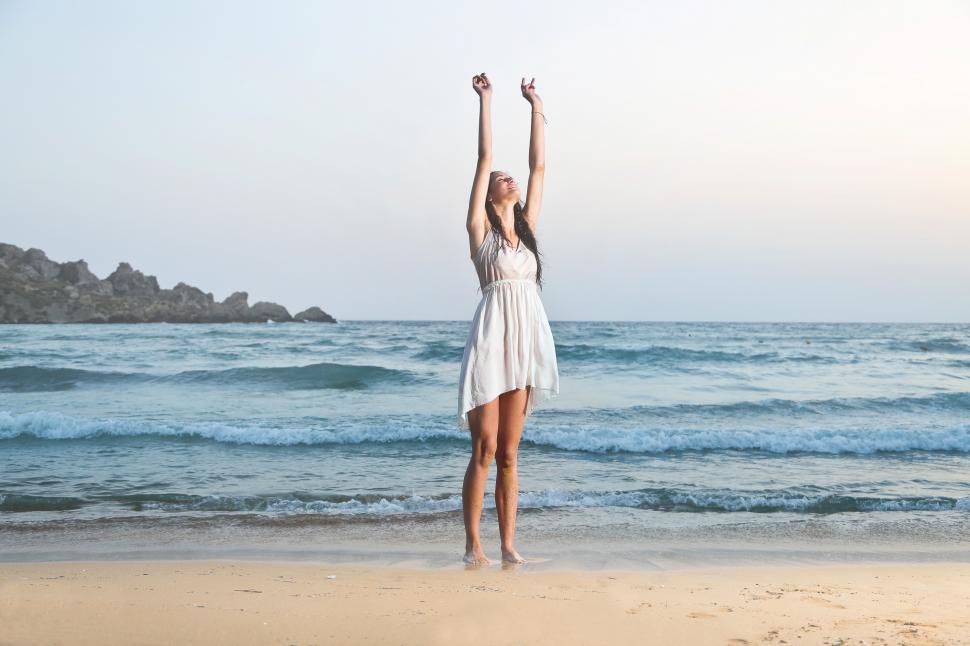 Free Image of A young brunette woman standing on the beach with her hands up 