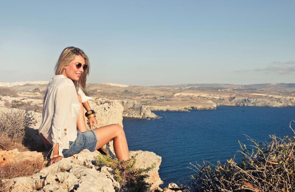 Free Image of A young blonde wearing shorts sitting on a rock by the sea 