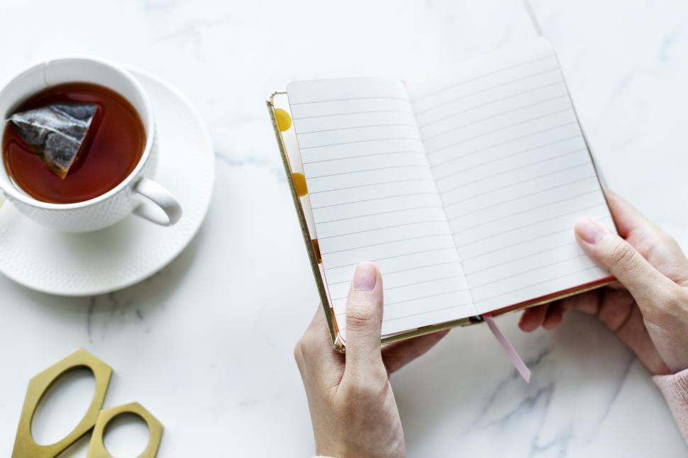 Free Image of Overhead view of a person s hands holding a notebook surrounded with a cup of tea and scissors on the marble surface 