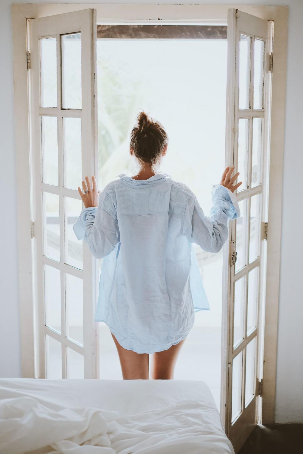 Free Image of Backside View of woman in blue shirt standing near bedroom window 