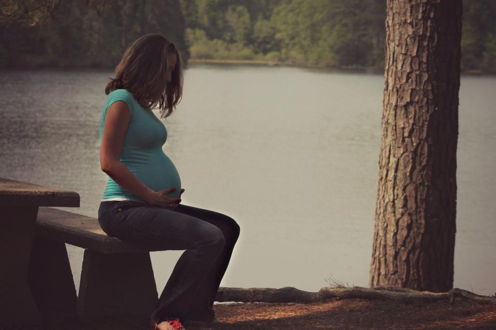 Free Image of Pregnant Woman touching belly on bench with lake in the background  