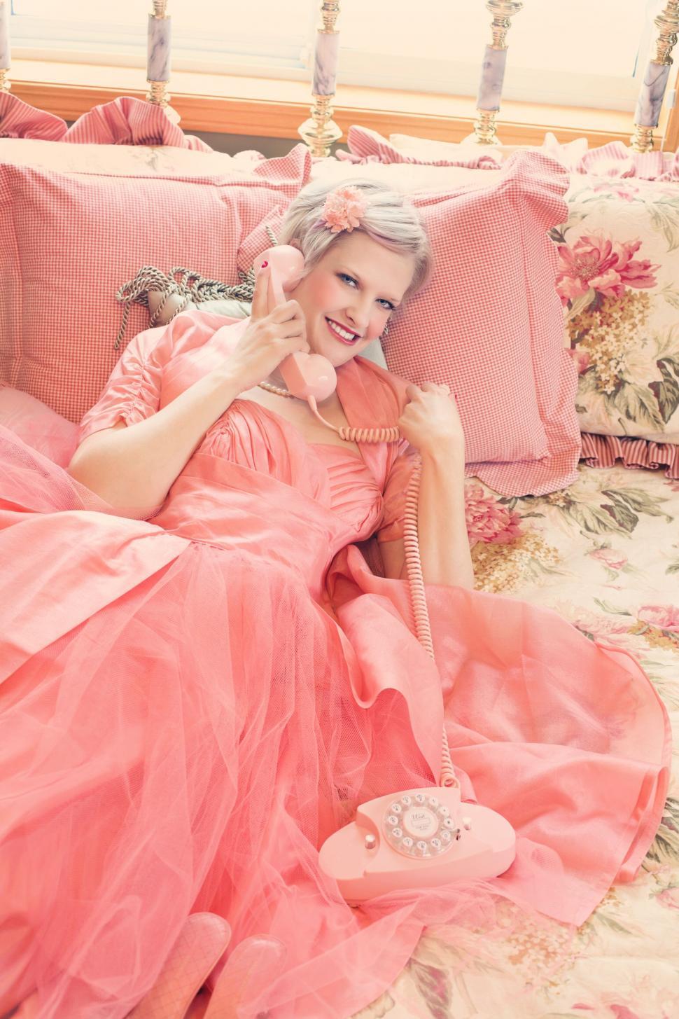 Free Image of Smiling Blonde Woman Talking on pink rotary dial telephone in bedroom 
