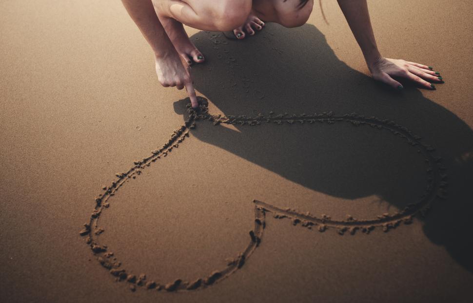 Free Image of A young woman at the beach hand drawing a heart in the sand 