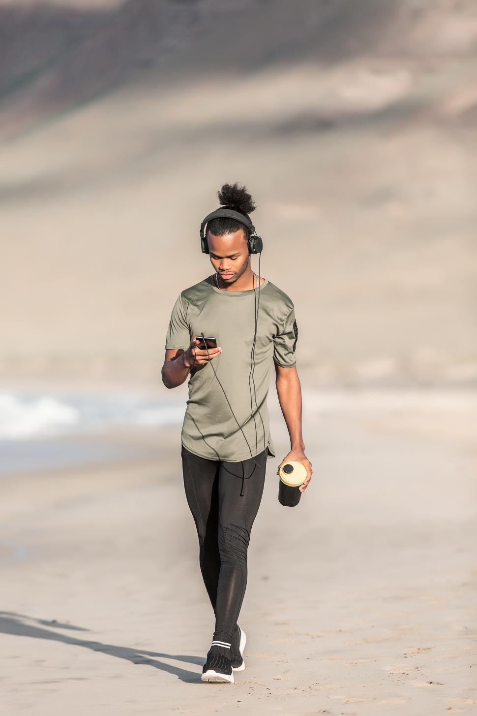 Free Image of Black guy with cup and smartphone walking on seashore 