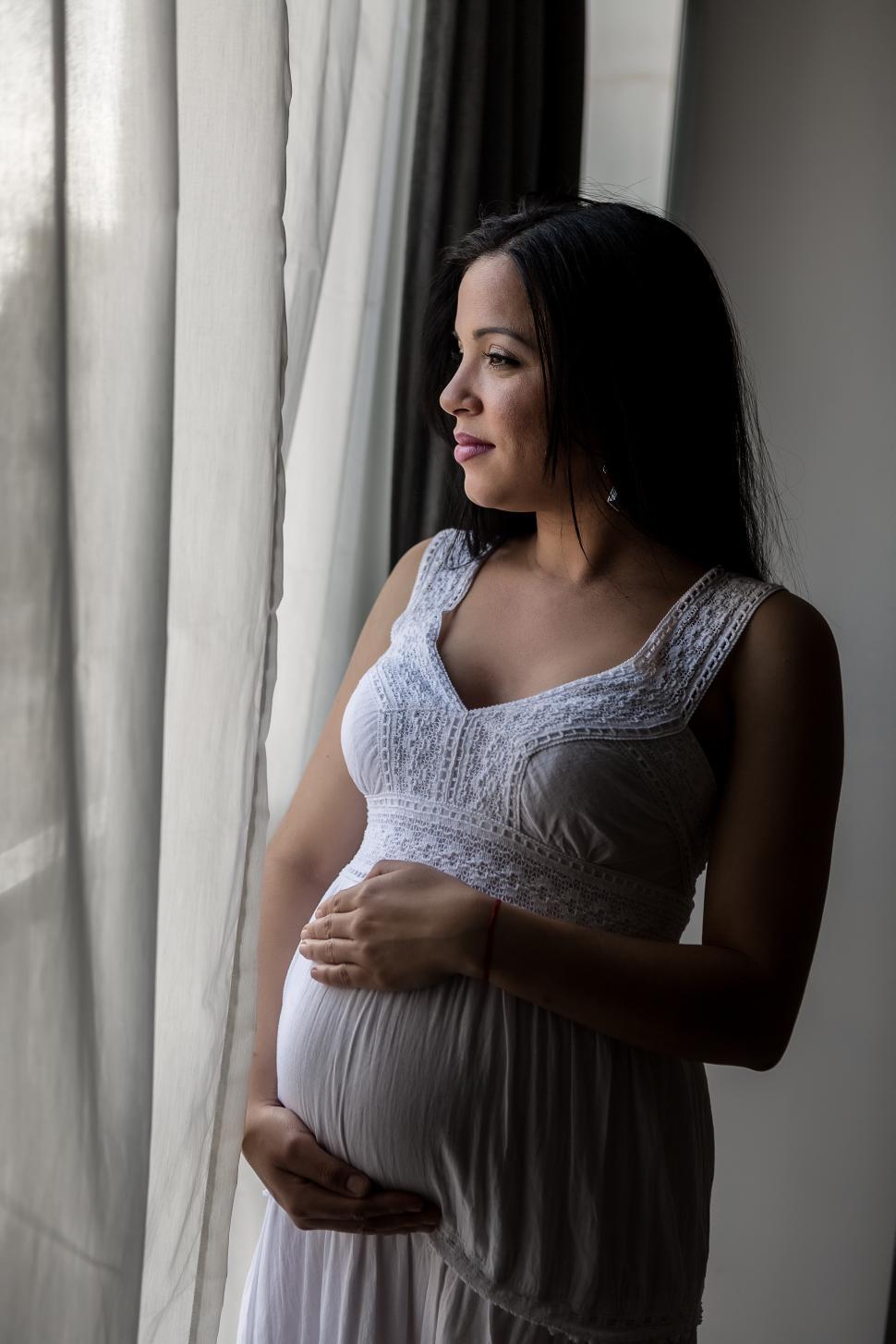 Free Image of Pregnant woman looking out window, hands on belly 