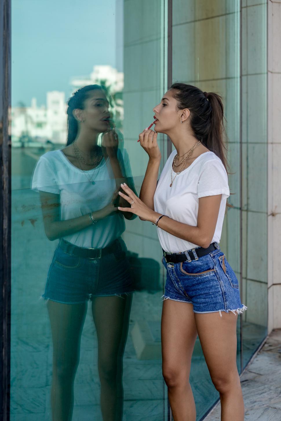 Free Image of Slim woman applying lipstick near glass wall 