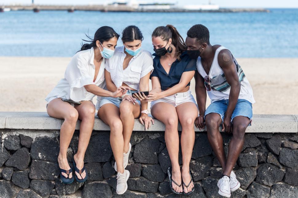 Free Image of Group of friends in masks using smartphone on seashore 