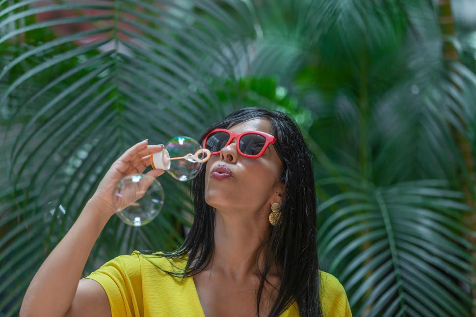 Free Image of Woman in yellow dress and sunglasses with red frames blowing bubbles 