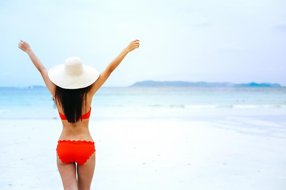 Free Image of Woman in a bikini stands on the beach with her arms raised 