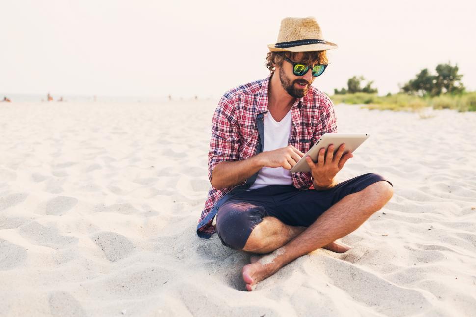 Free Image of Handsome cheerful hipster man with beard in straw hat, checkered shirt, with tablet device on the beach 