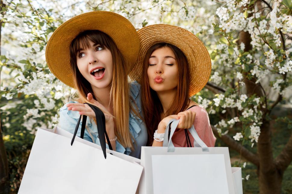 Free Image of Two stylish happy women with white shopping bags posing on blooming trees background 