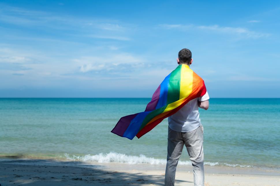 Free Image of Young gay man wrapped in pride rainbow flag while standing on the beach 