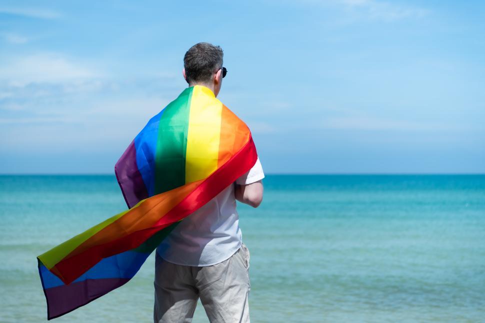 Free Image of Young gay man covered in rainbow flag, looking at the ocean 