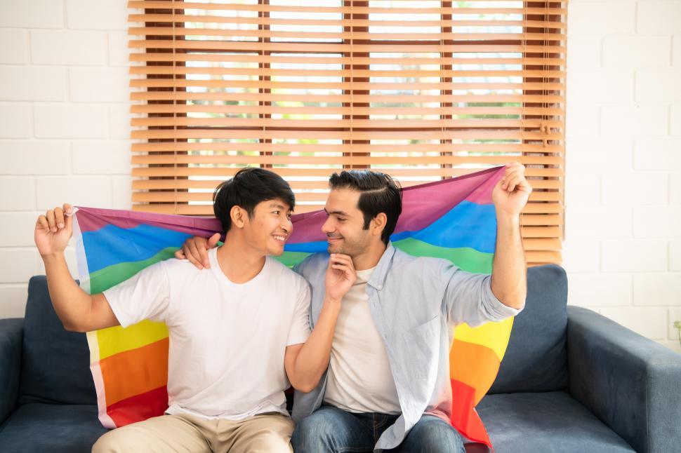 Free Image of LGBT couple - two men sitting on the couch together holding pride flag arond themselves 