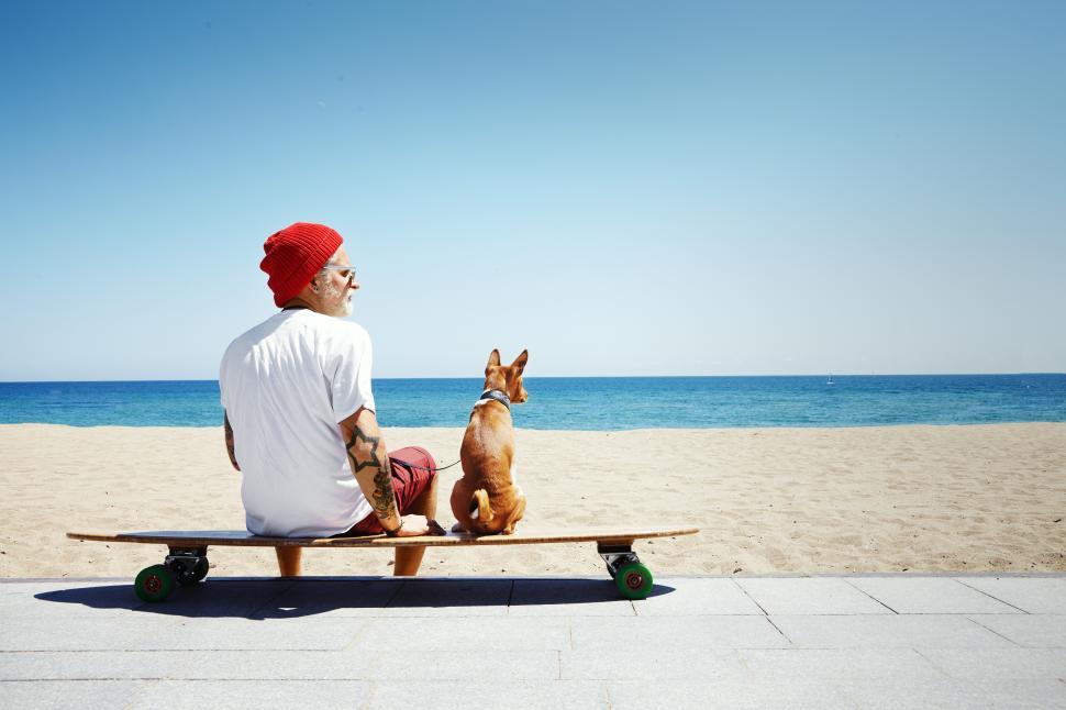 Free Image of Bearded man sitting on longboard with his dog, looking at the ocean 