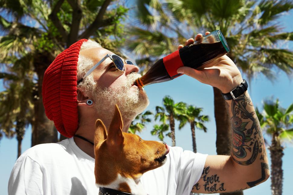 Free Image of Man drinks a soda while his dog looks at him, palms in the background 