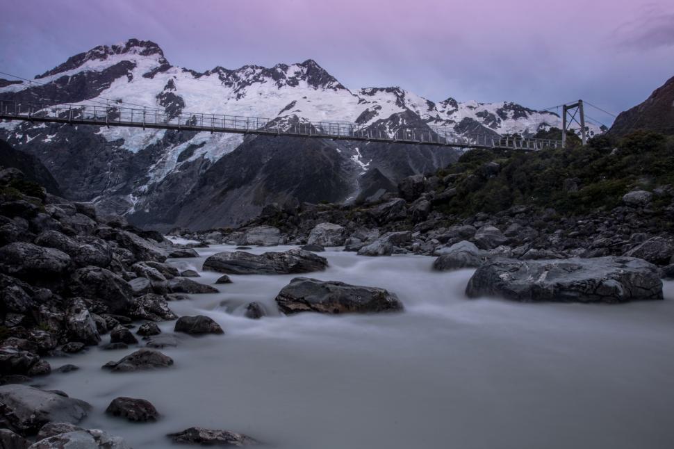 Free Image of A bridge over a river with rocks and snow covered mountains 