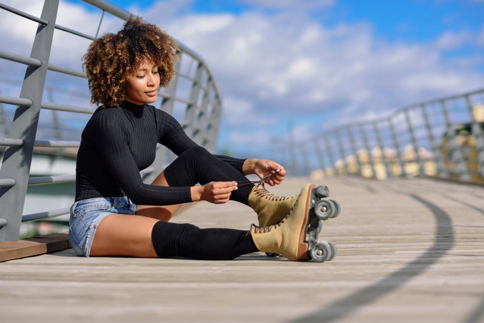 Free Image of Young smiling black girl sitting on urban bridge and puts on skates. 