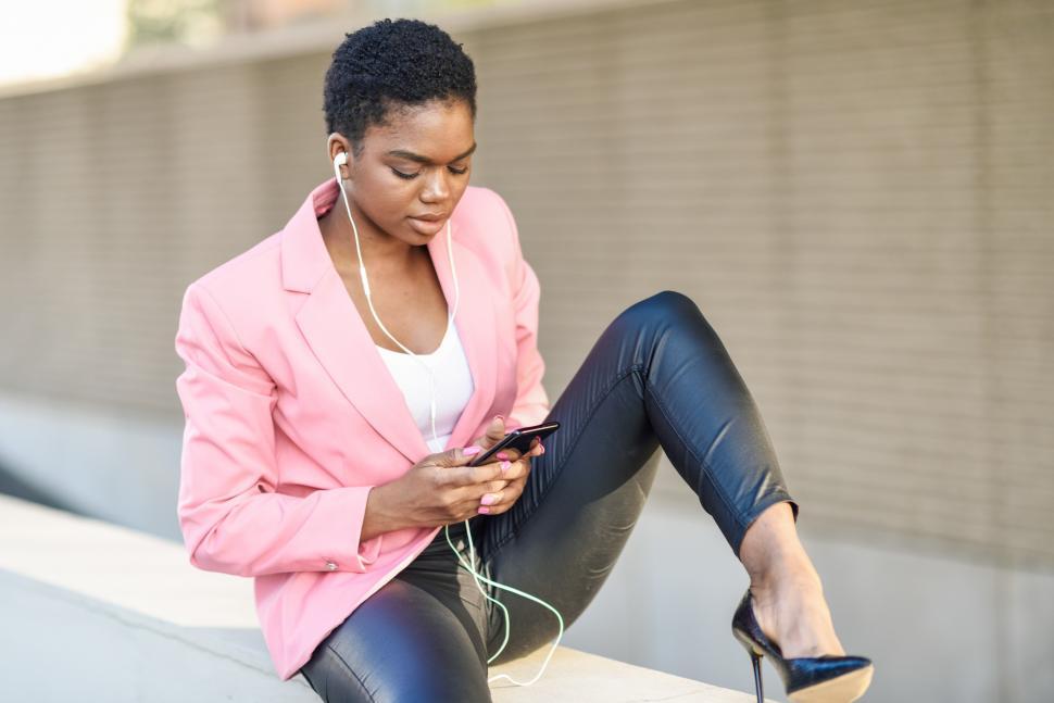 Free Image of Black businesswoman sitting outdoors using smartphone with earphones 