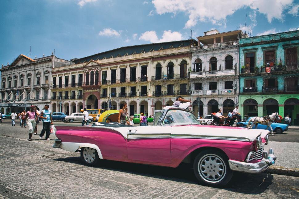 Free Image of A pink and white convertible car parked on a street with buildings and people 