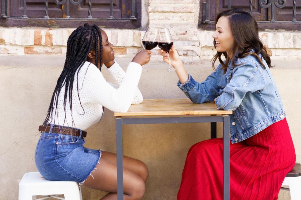 Free Image of Two women making a toast with red wine sitting at a table outside a bar. 