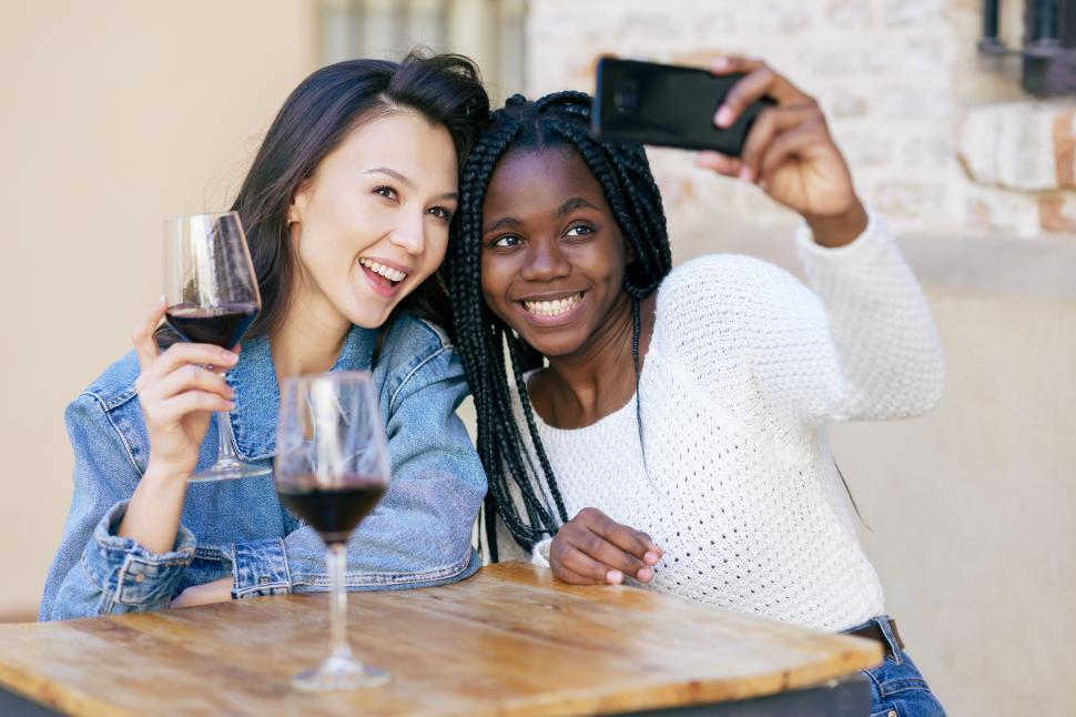 Free Image of Two friends making a selfie sitting at a table outside a bar while drinking a glass of red wine. 