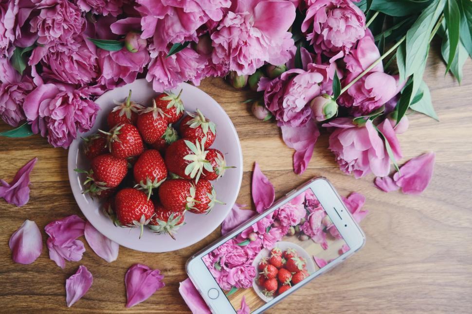 Free Image of A bowl of strawberries and a phone next to pink flowers 