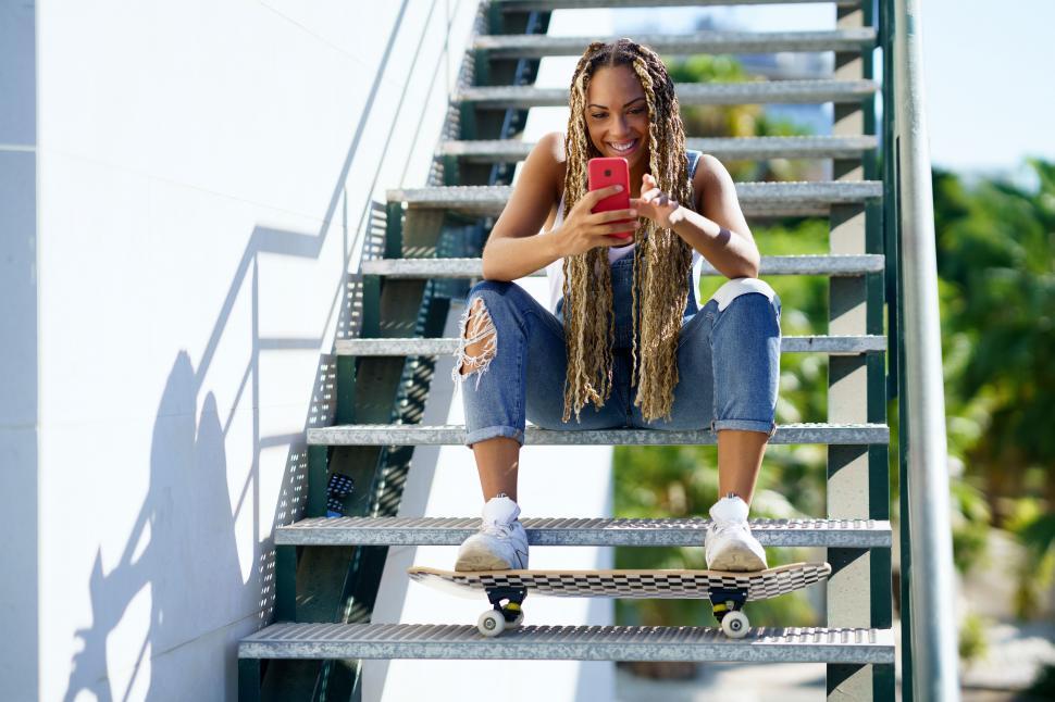Free Image of Black woman with coloured braids, consulting her smartphone with her feet resting on a skateboard. 