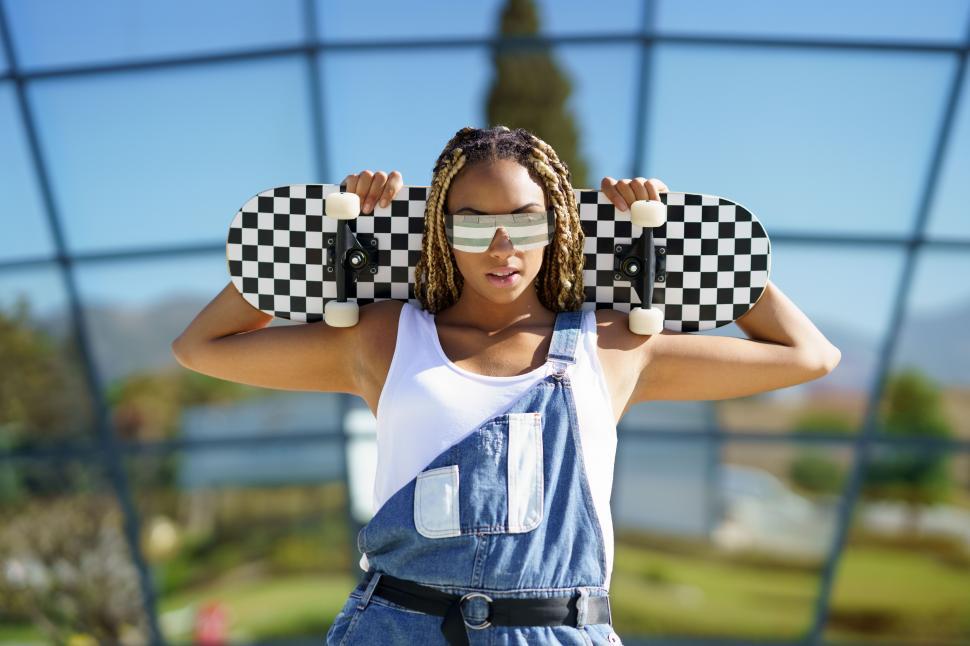 Free Image of Black woman dressed casual, looking at camera with modern sunglasses and a skateboard. 