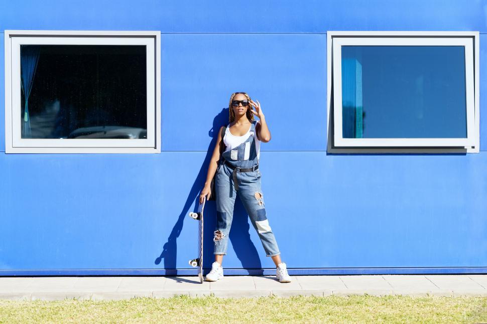 Free Image of Black woman dressed casual, with modern sunglasses and a skateboard on blue wall background. 
