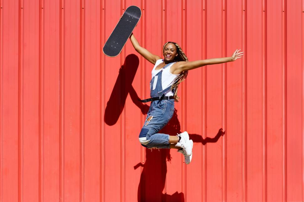 Free Image of Black woman dressed casual, wtih a skateboard jumping with happiness on red urban wall background. 